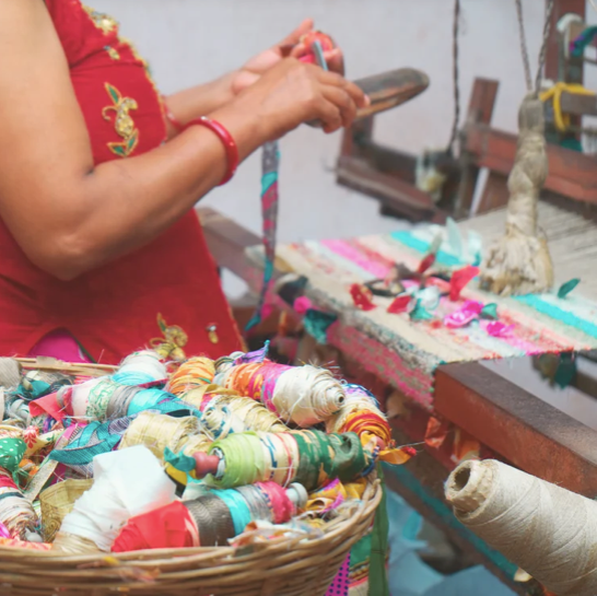 Weberin von Local Women's Handicrafts in Nepal am Handwebstuhl, davor ein Korb mit bunten Garnspulen